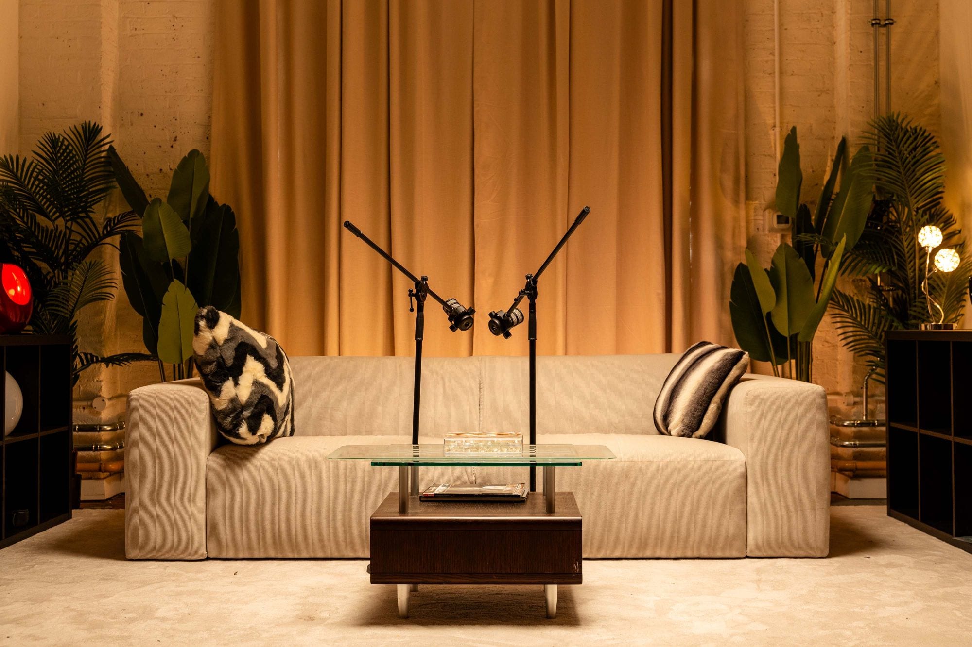 The cream corduroy sofa in the Creative Mike Studios room with two microphones on boom arms over the glass coffee table, gold curtains and palms behind