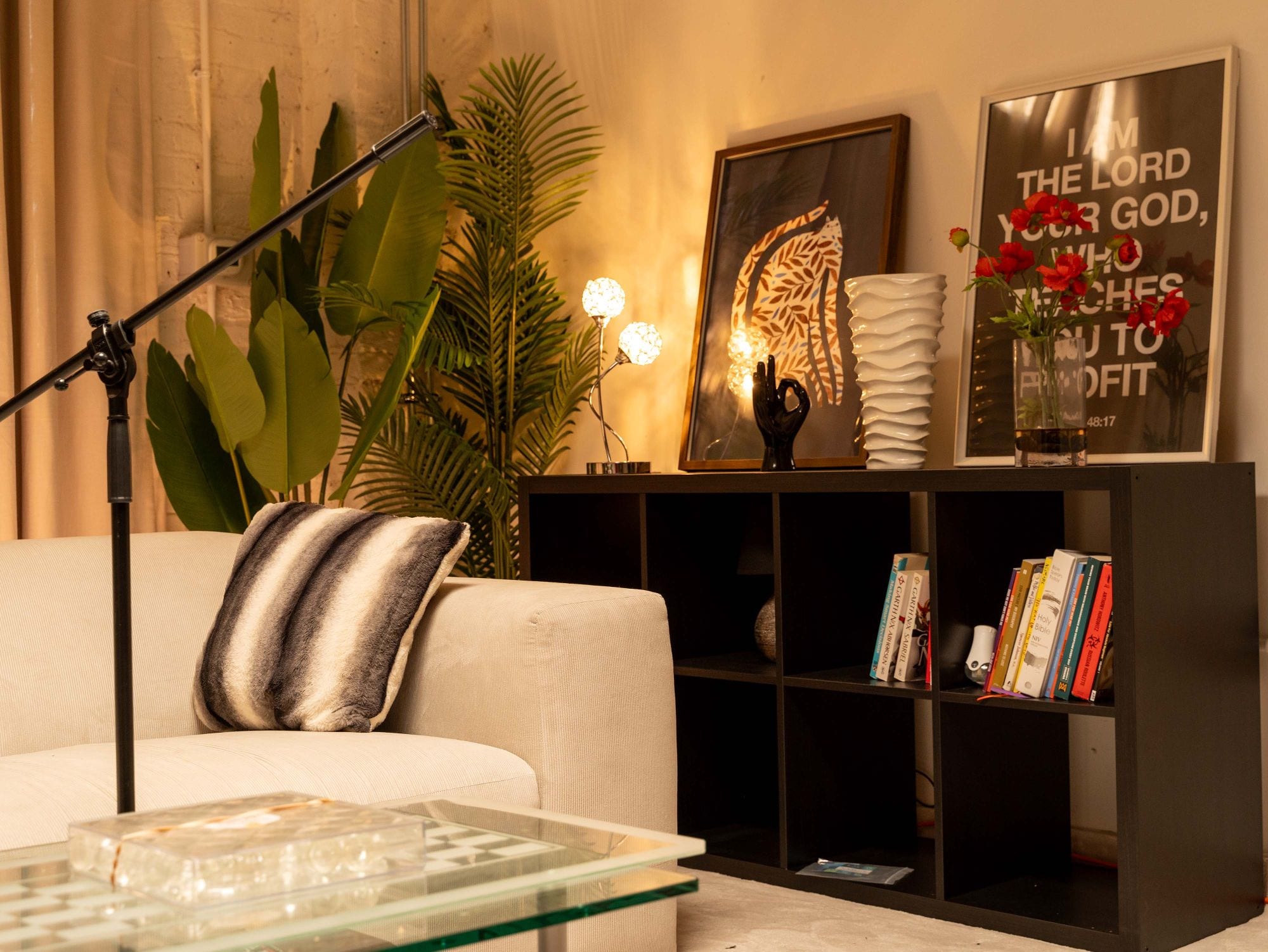 Corner of the room with a black cube shelf of books, a white sculpted vase, red flowers and two framed prints under warm lamplight
